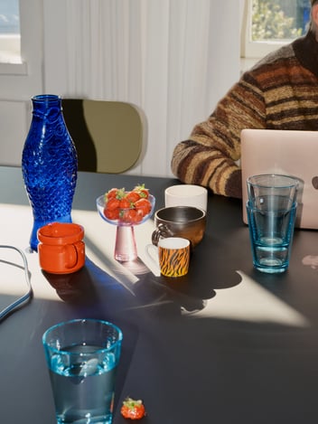 person behind a laptop, multiple random items on the table, e.g. a vase and a coffee cup.